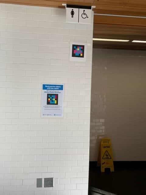 White tile wall next to the women's restroom at White Plains: above, the woman icon and the accessibility icon; in the middle, a NaviLens code framed in white; below, an official MTA blue sign reading «NaviLens for riders with low vision · We're testing NaviLens at White Plains Station»