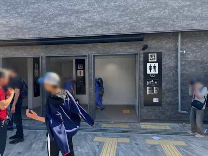 Toilet access “トイレ · Toilets” with men, women and accessible-toilet pictograms alongside a square NaviLens code, yellow tactile paving and visitors entering