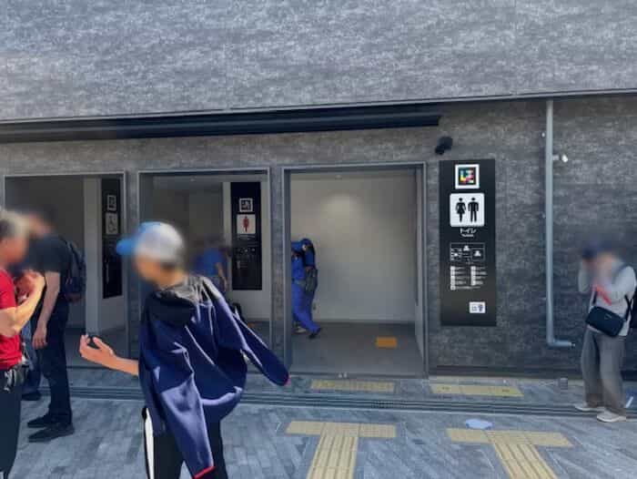 「トイレ · Toilets」入口の男性・女性・多目的トイレピクトグラムと並ぶ正方形のNaviLensコード、黄色い点字誘導ブロックと入る来場者たち