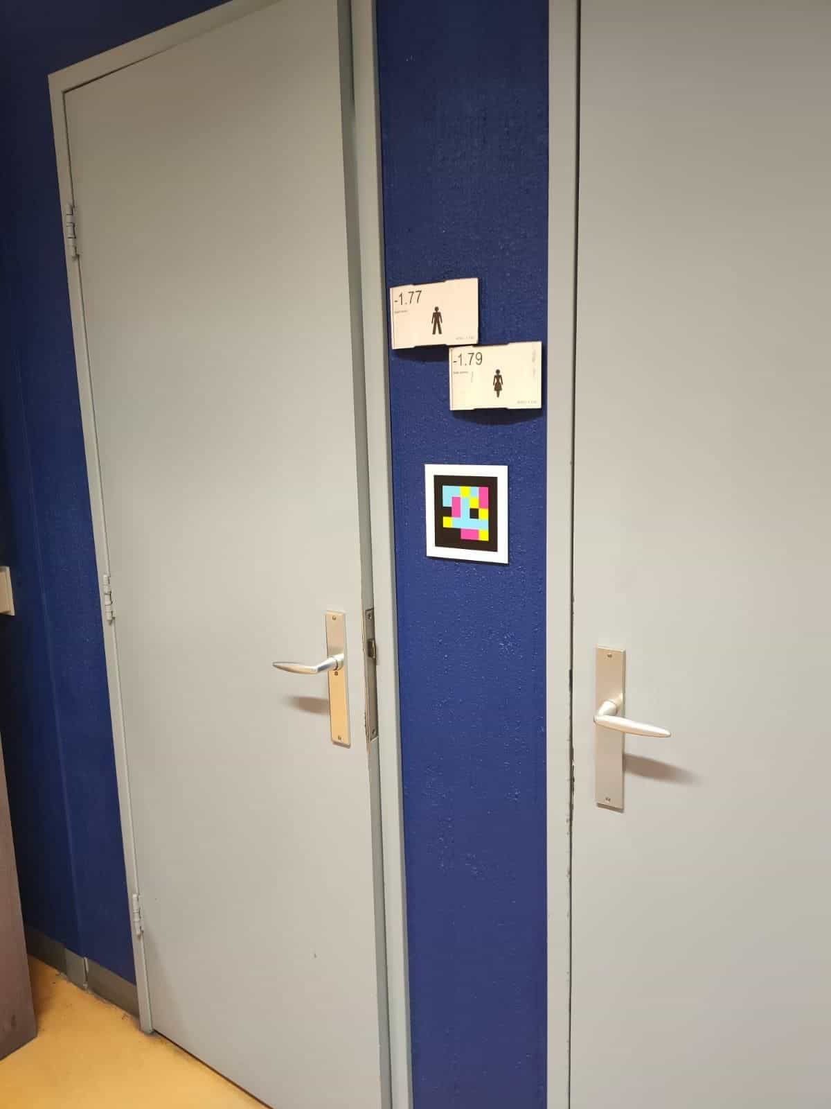 Zwei graue Toilettentüren des Radboudumc mit blauem Rahmen. An der Mittelsäule zwei kleine weiße Schilder «-1.77 Heren» und «-1.79 Dames» mit Piktogrammen; darunter ein weiß umrahmter NaviLens-Code, der in 39 Sprachen ansagt, welche Tür zu welcher Toilette führt