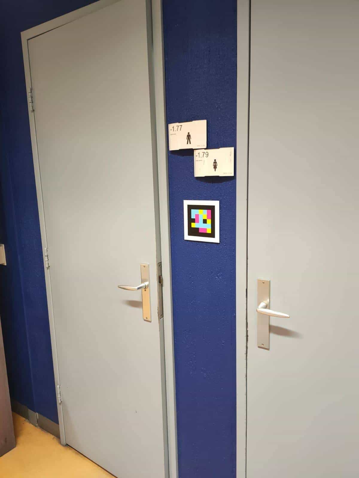 Two grey toilet doors at Radboudumc with blue frames. On the centre column, two small white signs "-1.77 Heren" and "-1.79 Dames" with their pictograms; below, a white-framed NaviLens code that announces in 42 languages which door leads to which toilet