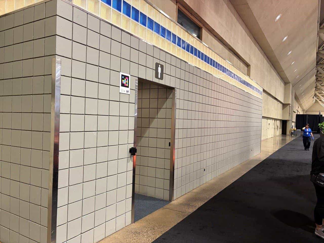 A corridor in the Metro Toronto Convention Centre hall, with a pale tiled wall topped by a band of blue and amber glass block. Beside the toilet entrance, marked with its pictogram, a NaviLens code with the TIFF logo.