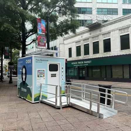 Throne unit on a downtown Atlanta sidewalk, in front of the Azalea Fresh Market building, with a side sign reading «Free Restroom / Baño Gratis» and a NaviLens code next to the entrance.