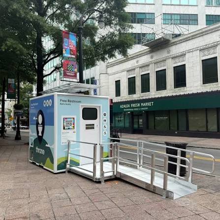 Cabina Throne en una acera del centro de Atlanta, frente al edificio del Azalea Fresh Market, con el cartel lateral «Free Restroom / Baño Gratis» y un código NaviLens junto a la entrada.