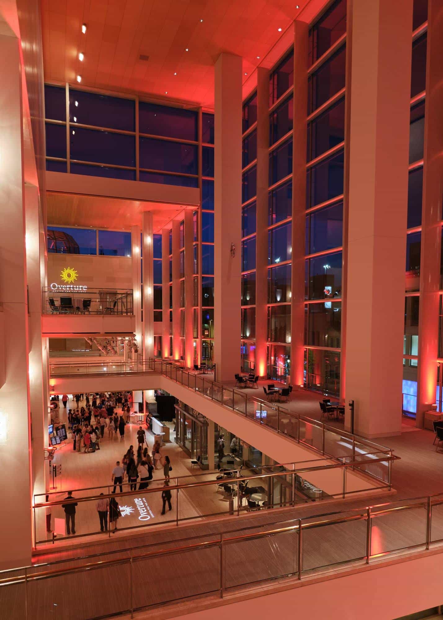 Overture Center atrium lit in red after a performance: the audience fills the lower level beneath the Overture logo projected on the floor and the wall, with the upper-level walkways in the foreground