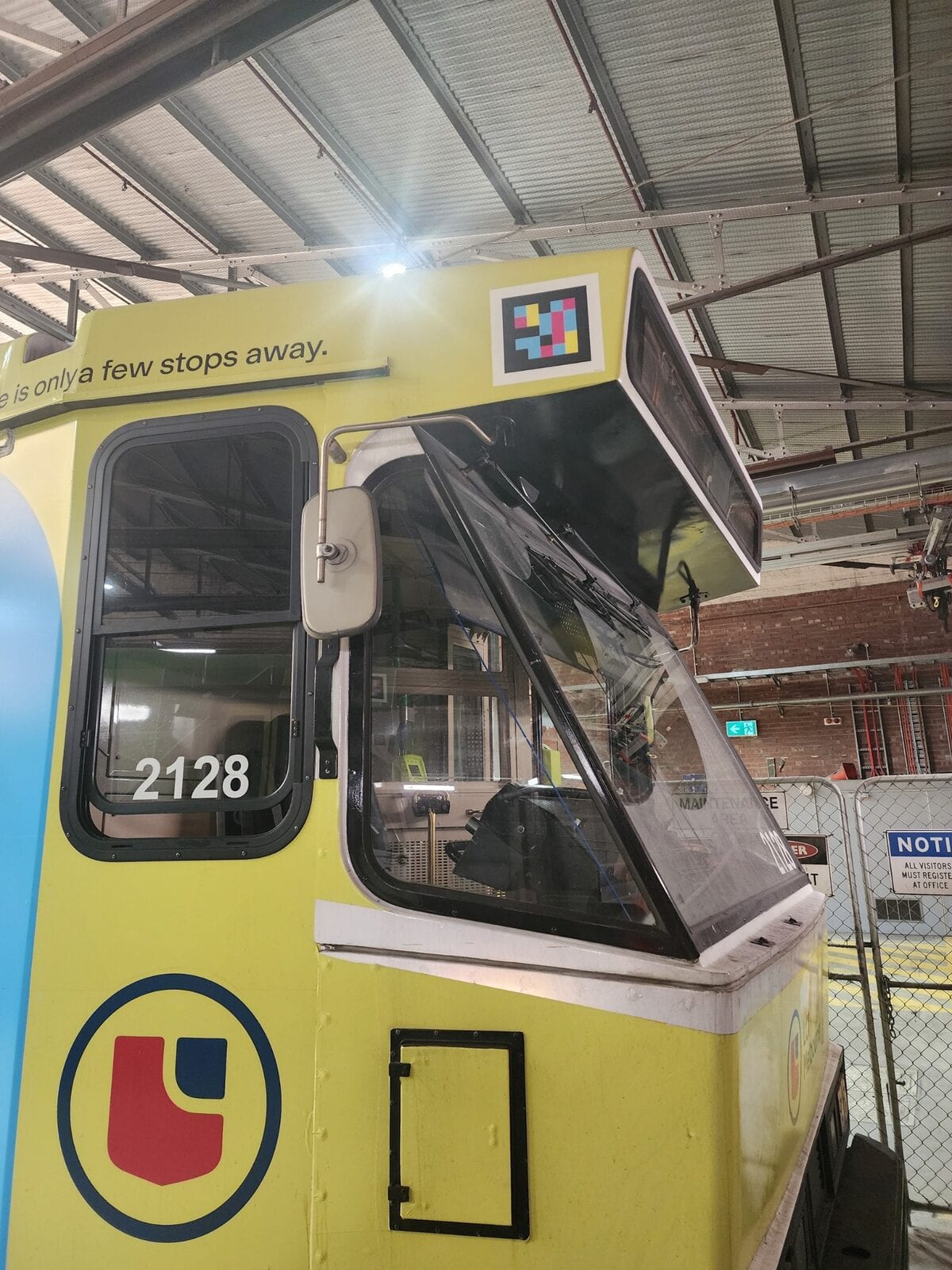 Cab of yellow B-Class tram 2128 at Yarra Trams’ Preston Workshops depot, corrugated roof behind; on the white front, next to the text «is only a few stops away.», a large white-framed NaviLens code; the historic blue-and-red «MMTB» logo on the driver’s door and the black maintenance hatch at the base of the nose