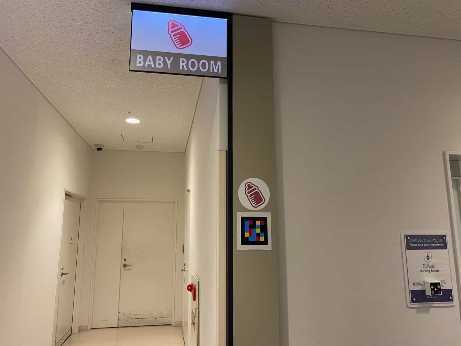 Entrance to the “BABY ROOM · Nursing Room” at Kobe Airport with illuminated sign, baby-bottle pictogram and NaviLens tag