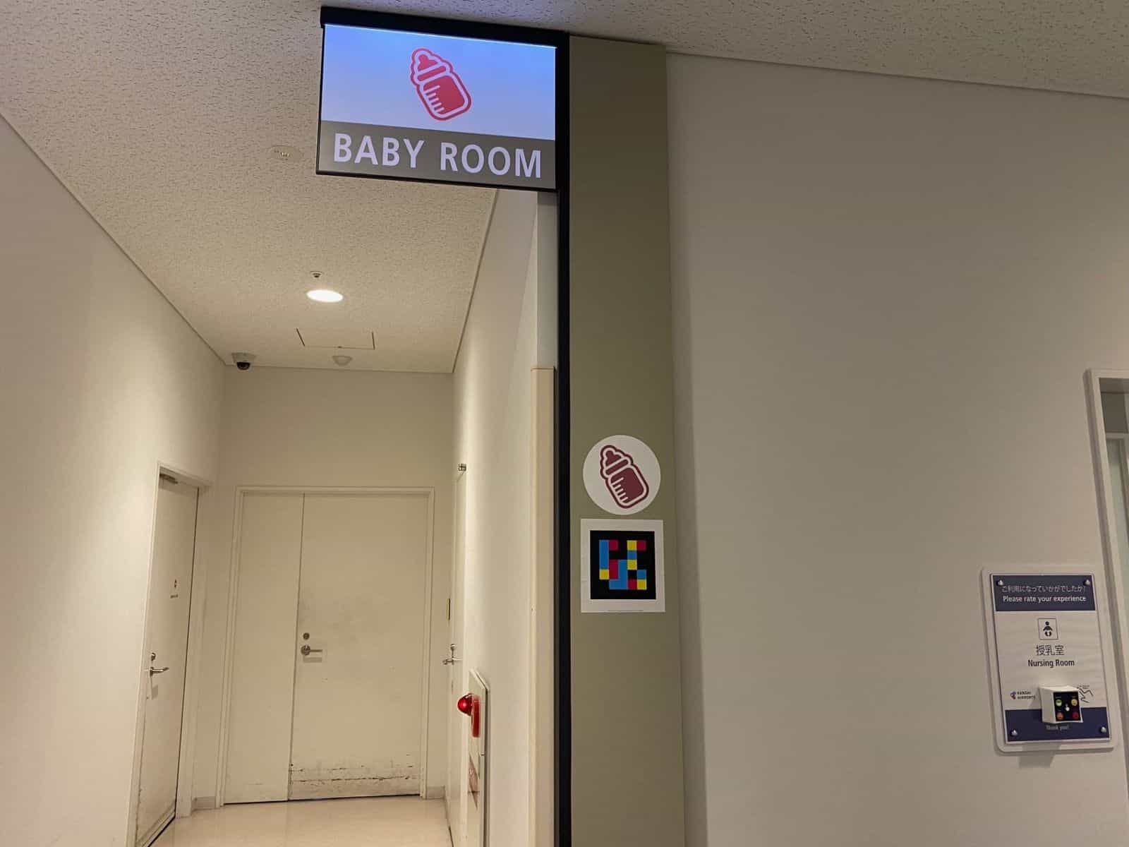 Eingang zum «BABY ROOM · Nursing Room» im Kobe Airport mit Leuchtschild, Babyflaschen-Piktogramm und NaviLens-Tag