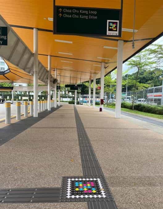 Bahía exterior de buses bajo marquesina amarilla: cartel direccional negro colgado del techo con flechas hacia «Choa Chu Kang Loop» y «Choa Chu Kang Drive» y un código NaviLens impreso a la derecha del cartel; en primer plano, suelo con franja podotáctil y un código NaviLens grande sobre baldosa blanca al borde de la zona de espera