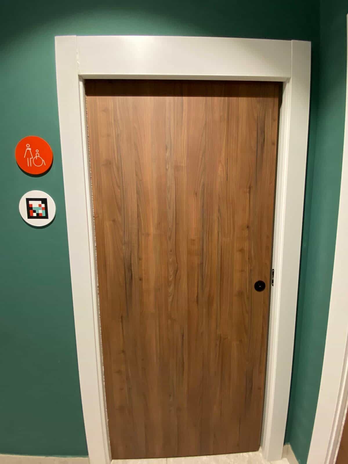 Tür eines Gemeinschaftsbads am ibis Styles Sevilla im grünen Flur: hölzerne Schiebetür und an der Wand ein oranger Disc mit Piktogramm für barrierefreies WC und darunter NaviLens-Code auf weißem Disc