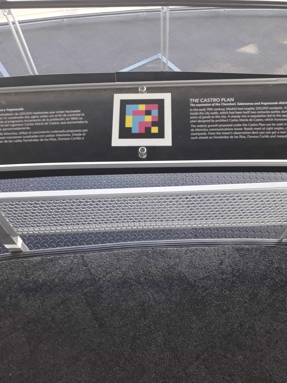 Barandilla informativa del mirador del Faro de Moncloa: sobre un panel negro que explica «The Castro Plan» en español e inglés, un código NaviLens cuadrado a colores enmarcado en blanco permite escuchar el contenido del panel a través del móvil