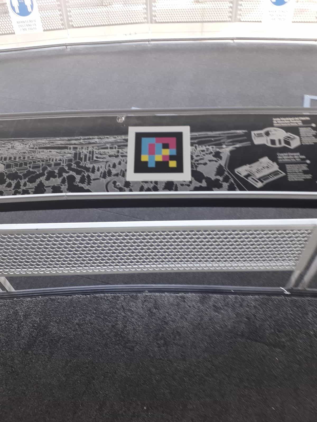 Detalle de un panel de la barandilla informativa del Faro de Moncloa con un mapa aéreo grabado en relieve sobre fondo negro; en el centro, un código NaviLens cuadrado a colores enmarcado en blanco permite escuchar la descripción del panorama