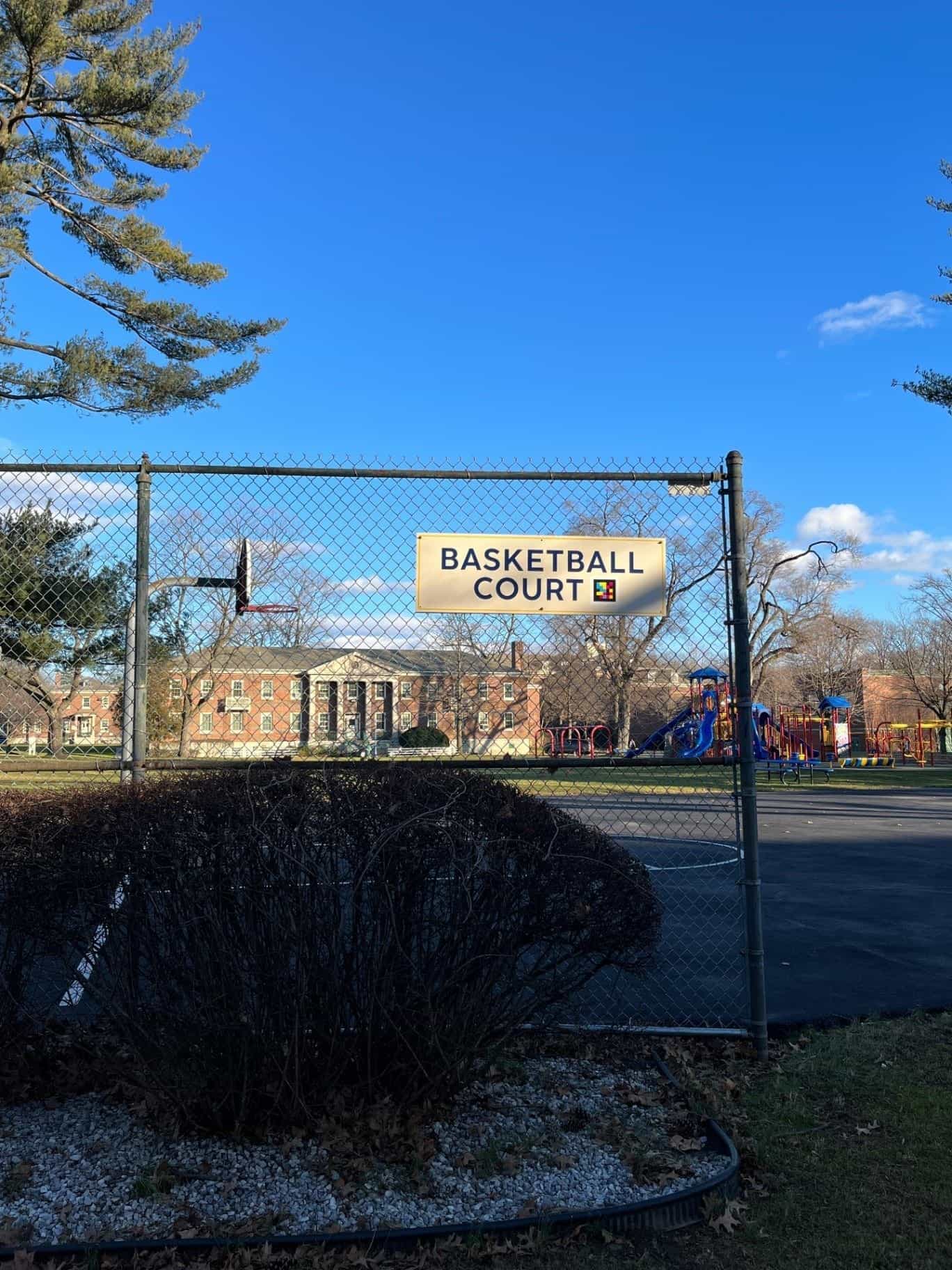 金属フェンス越しに見えるNYISEのバスケットボールコート:「BASKETBALL COURT」と書かれたクリーム色とターコイズ色の看板があり、スポーツ施設を読み上げるNaviLensコードが設置されている