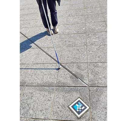 Person mit weißem Blindenstock geht auf einen NaviLens-Code zu, der auf dem grauen Granitpflaster vor dem Museum klebt