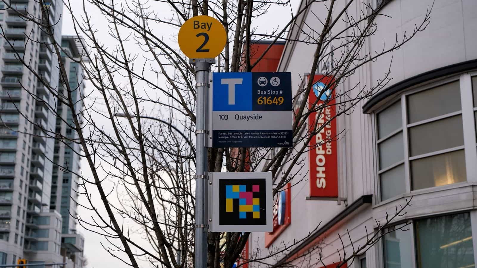 Palo di fermata TransLink a New Westminster: disco giallo «Bay 2» nella parte alta, sotto un cartello azzurro e bianco con la T del trasporto pubblico, «Bus Stop # 61649» in arancione e la linea «103 Quayside»; sotto il cartello, una placca quadrata con un codice NaviLens a quadretti colorati su fondo nero; in fondo, edifici residenziali e la facciata rossa di Shoppers Drug Mart