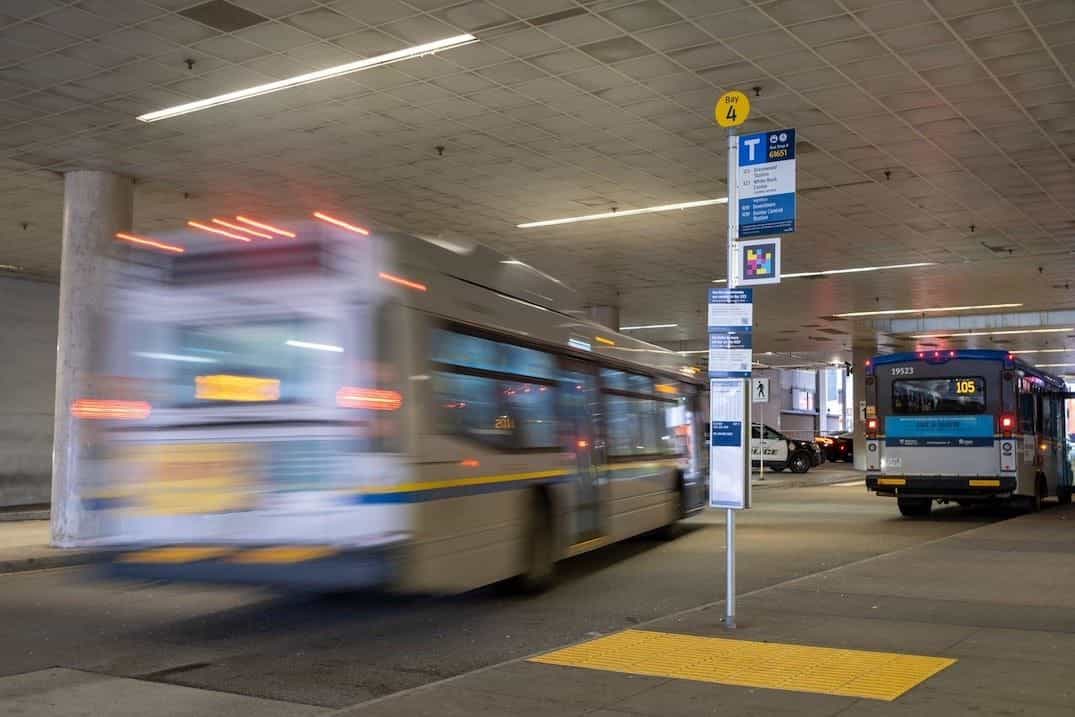 Interior cubierto de la estación de SkyTrain New Westminster: poste con disco amarillo «Bay 4», cartel azul «Bus Stop # 69651» con líneas 123/321 y N19, código NaviLens a colores debajo y bandas podotáctiles amarillas en el suelo