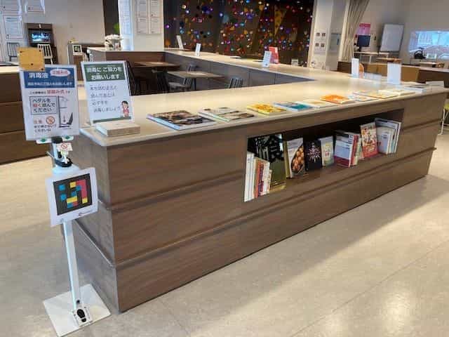 ビジョンパークの図書スペースと休憩エリア。料理本、木製カウンター、足元にNaviLensタグを貼ったアルコール消毒ディスペンサー、奥にはカラフルなキッズ・クライミングウォール