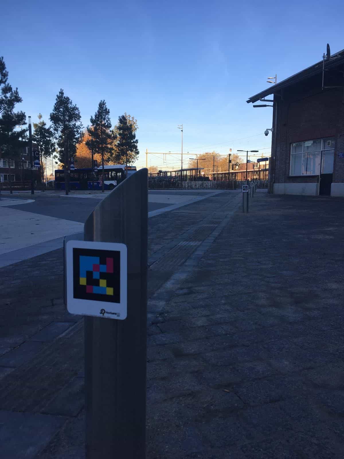 Dissuasore metallico curvo con codice NaviLens all’accesso delle banchine della stazione degli autobus di Roermond, con un autobus azzurro di Arriva e la stazione NS in fondo