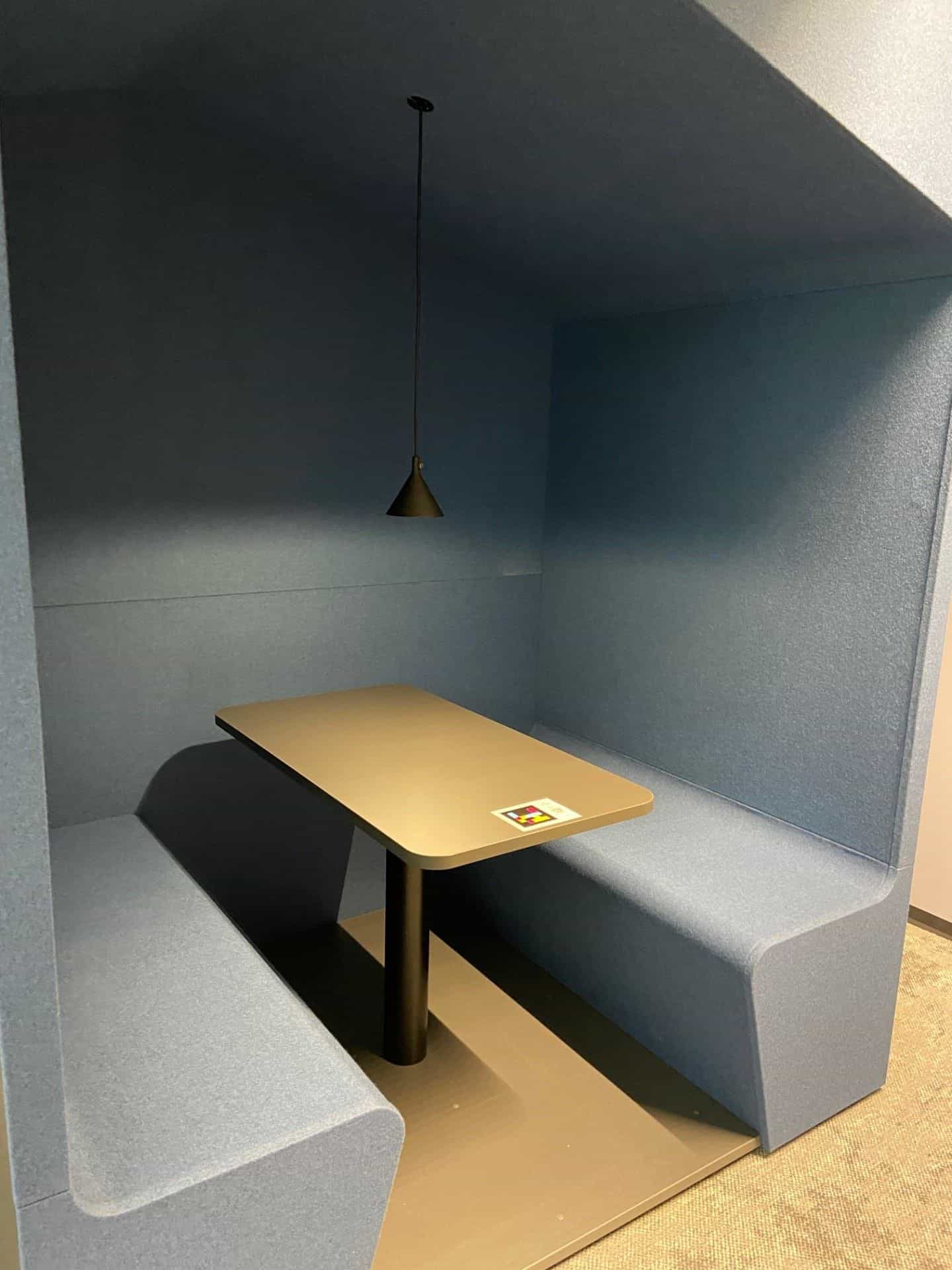 Booth acustico blu per 2-4 persone nella sede Bayer di Leverkusen — cartello NaviLens sul tavolo in legno con lampada a sospensione