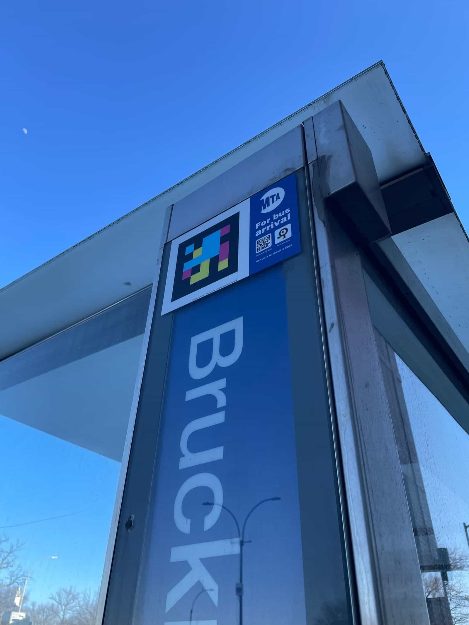 Dettaglio dal basso di una fermata su Bruckner Blvd con codice NaviLens ufficiale accanto alla targa azzurra della MTA «For bus arrival» nell’angolo superiore della pensilina