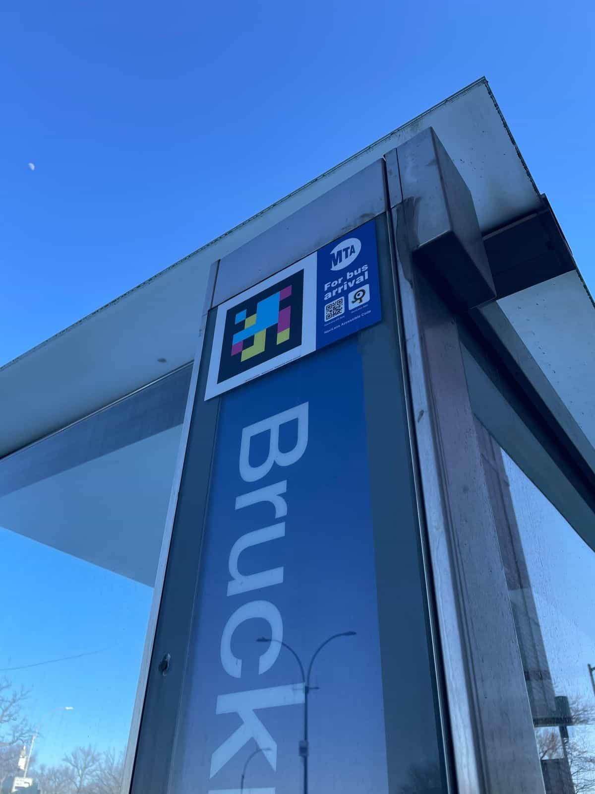 Detalle en contrapicado de una parada de Bruckner Blvd con código NaviLens oficial junto a la placa azul de MTA «For bus arrival» en la esquina superior de la marquesina
