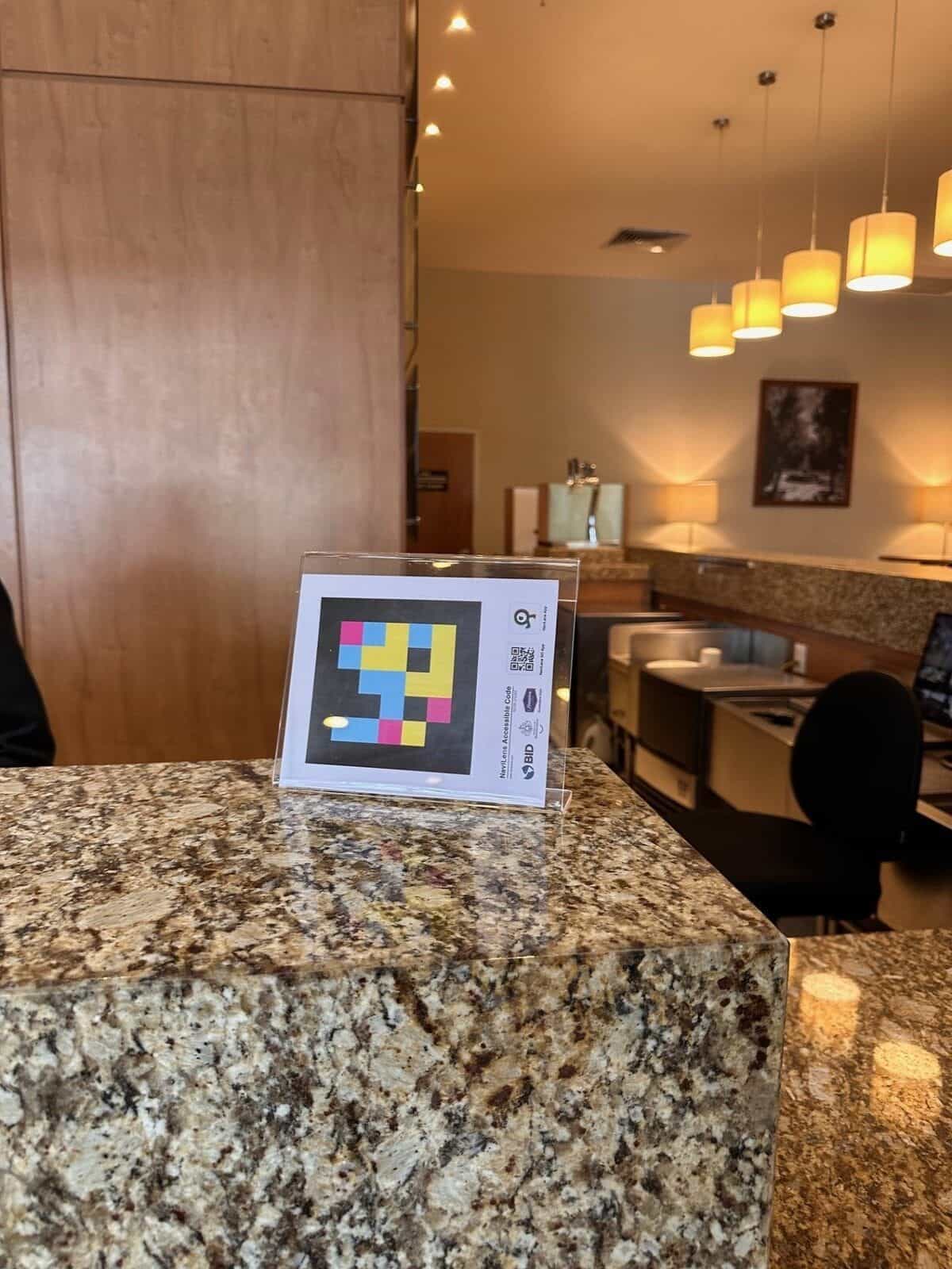 Panneau NaviLens sur le comptoir en granit du business center de l'Hampton by Hilton Guadalajara Expo, avec des suspensions en arrière-plan
