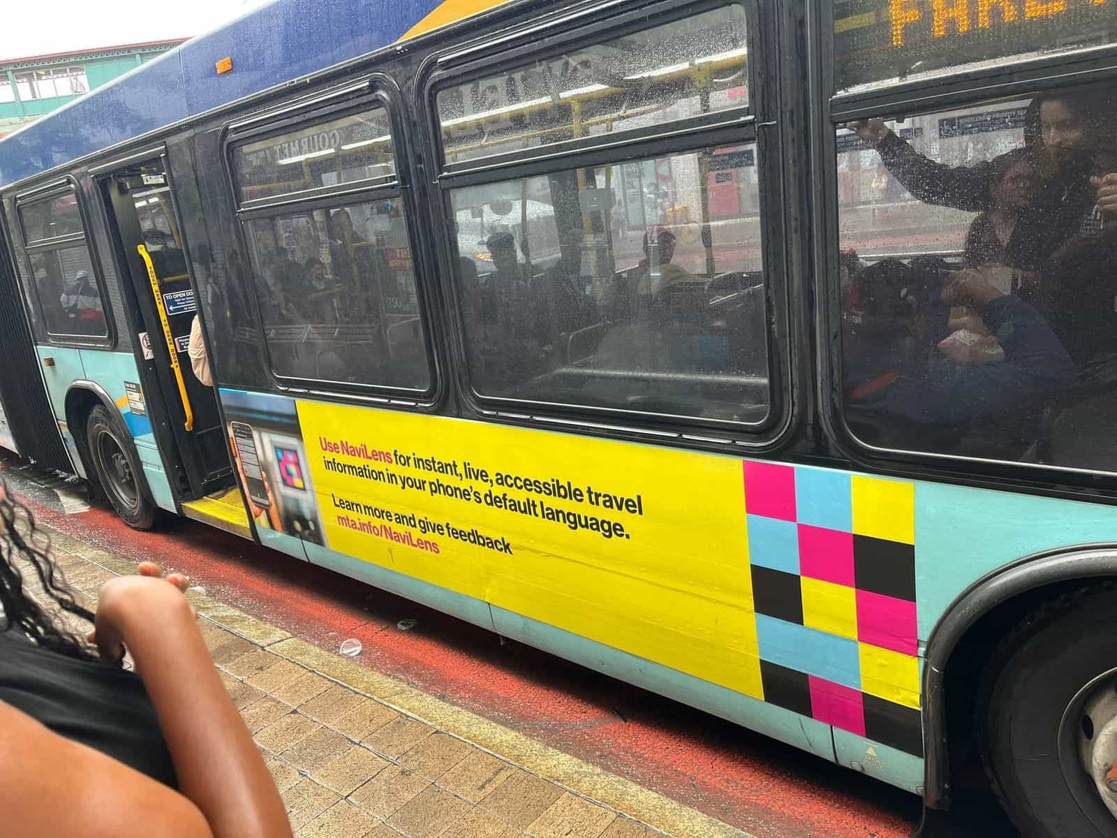 Bus Bx12 5558 fermo dietro la pensilina con codice NaviLens; fila di viaggiatori che entra dalla porta anteriore, etichetta OMNY a destra e codice NaviLens anche sul fronte del bus