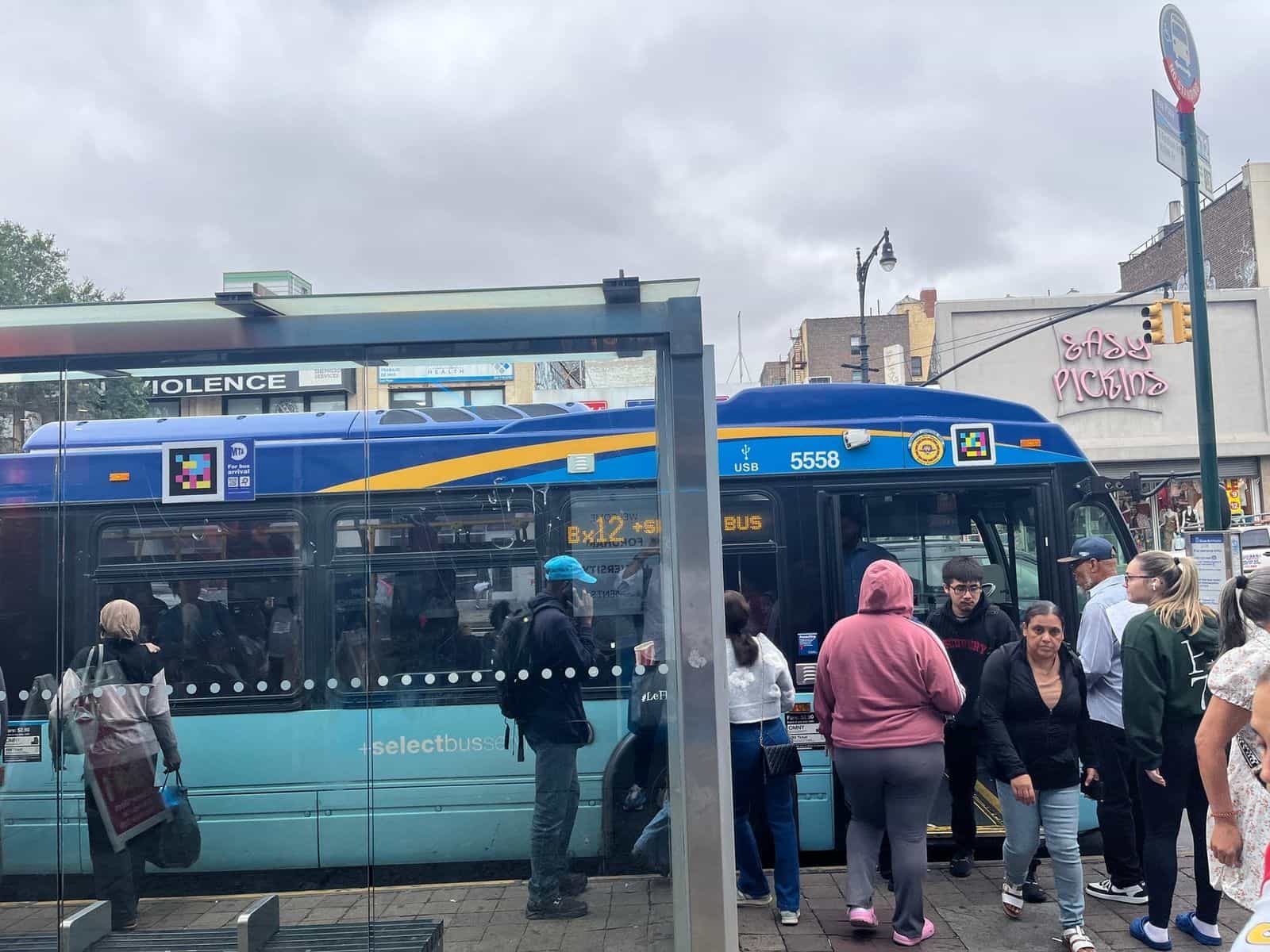 Bus MTA Bx12 +SELECT BUS numero 5558 fermo alla pensilina di Bay Plaza, con codici NaviLens visibili sul frontale del bus e sulla pensilina; viaggiatori che salgono dalla porta anteriore
