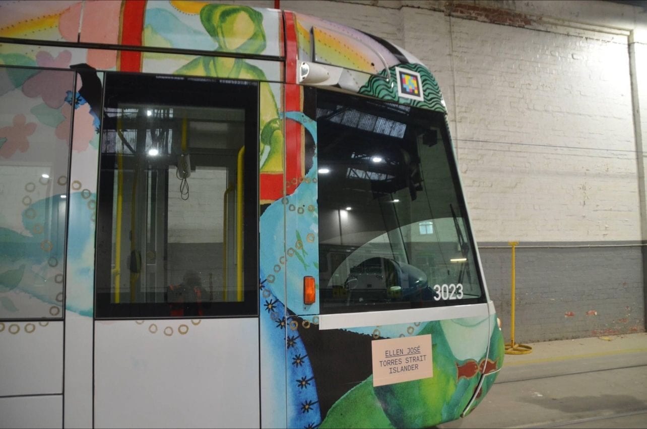 Front of Alstom C-Class tram 3023, wrapped as an Art Tram with artwork by Ellen José (Torres Strait Islander): green, blue and red watercolour painting on a white background; a white-framed NaviLens code above the cab and a small plaque under the headlight reading «ELLEN JOSÉ — TORRES STRAIT ISLANDER»
