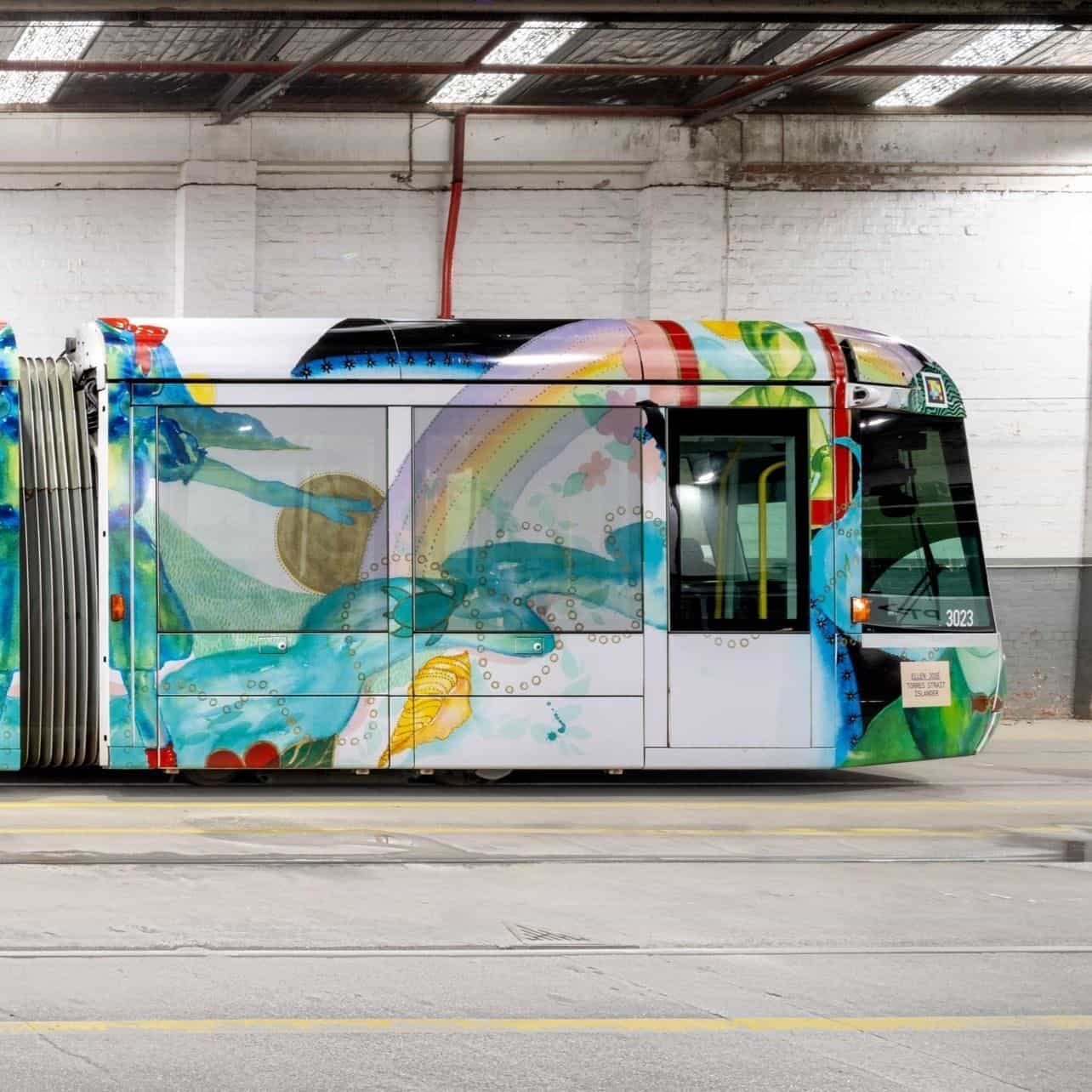 C-Class「Art Tram」3023 の側面ディテール。白い側面に水彩で描かれた虹と金色の太陽を持つ手、泡のような金色の円形モチーフ、ベルトラインの下に様式化された緑の魚。ノーズ上には NaviLens コード