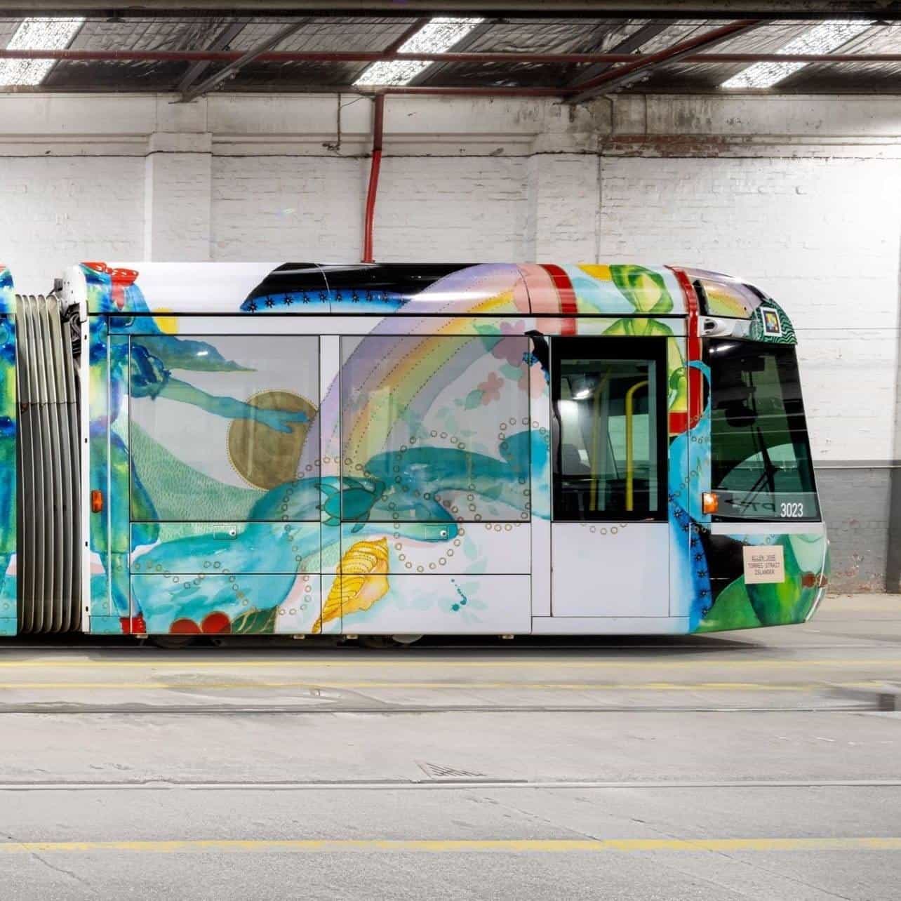 Side detail of C-Class “Art Tram” 3023: windows showing the illustration of a rainbow and hands holding a golden sun painted in watercolour on the white flank, circular golden bubble motifs and a stylised green fish under the waist line; the NaviLens code above the nose