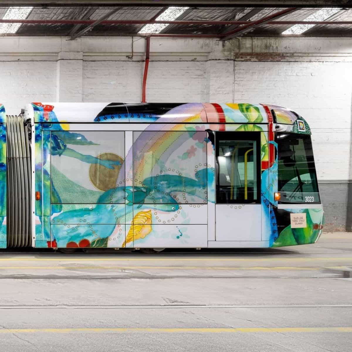 Detalle lateral del C-Class «Art Tram» 3023: ventanas con la ilustración de un arcoíris y manos que sostienen un sol dorado pintado en acuarela sobre el lateral blanco, motivos circulares dorados a modo de burbujas y pez verde estilizado bajo la línea de cintura; sobre el morro, el código NaviLens