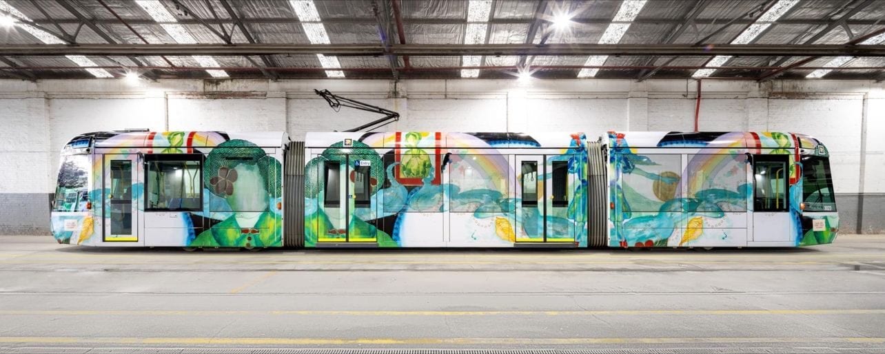 Full side view of C-Class 3023 «Art Tram» in the white-walled depot: five-module articulated car with Ellen José’s artwork wrapping it end to end —green and blue human figures, rainbow, fish, shells and botanical motifs— and the regulation yellow bands along the doors; a NaviLens code peeks above the right-hand cab