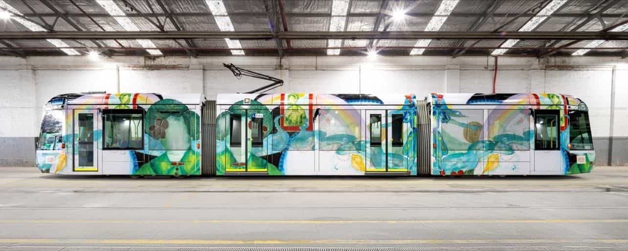 白い車庫内に置かれた C-Class 3023「Art Tram」の側面全景。5 連接の車体に Ellen José の作品が端から端まで巻かれ、緑と青の人物像、虹、魚、貝、植物モチーフが描かれ、ドア下部には規定の黄色帯。右側運転台の上に NaviLens コードが覗く