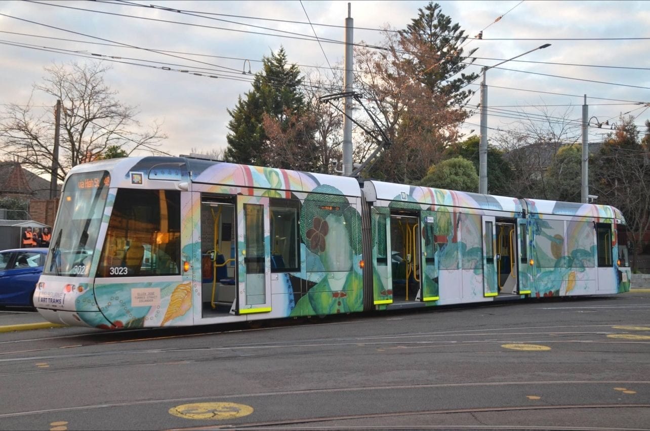 C-Class «Art Tram» 3023 with the Ellen José wrap in commercial service on a suburban Melbourne roundabout, with overhead wires, bare trees and the destination board «Via Hash St — 48»; the white-framed NaviLens code can be seen by the cab