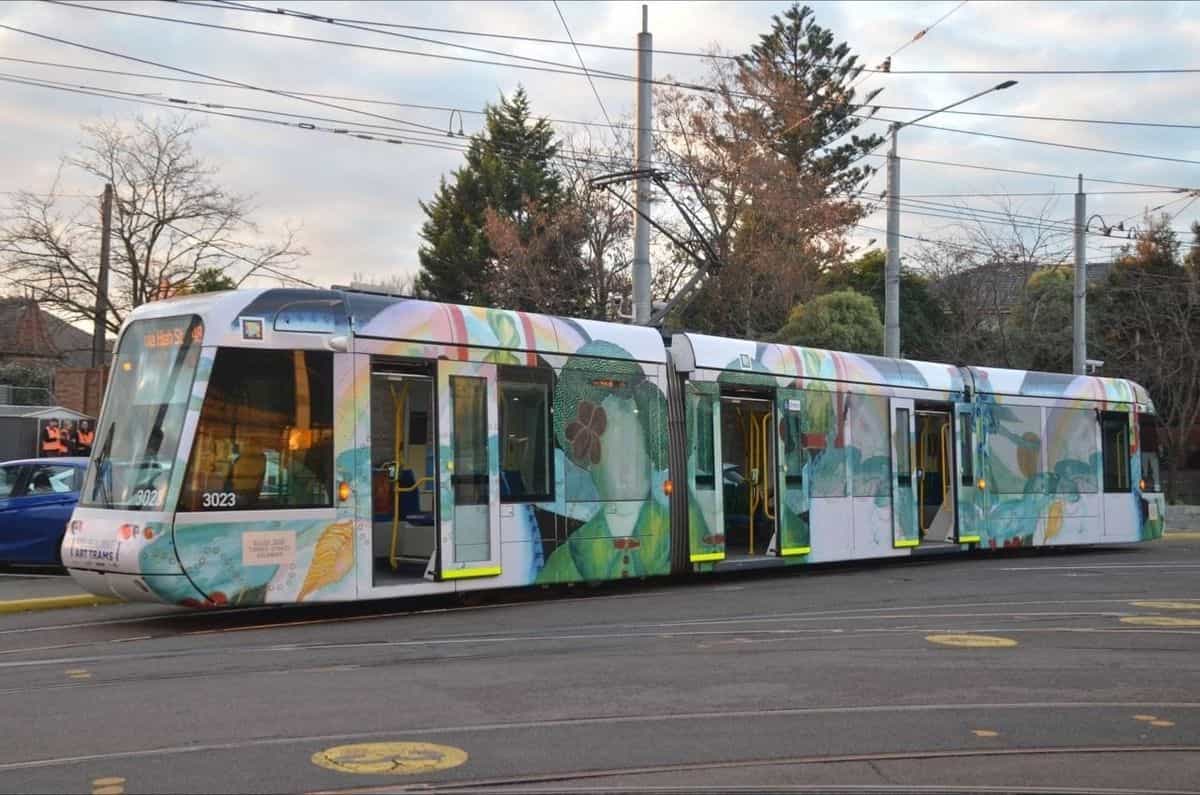 C-Class «Art Tram» 3023 con el wrap de Ellen José operando en servicio comercial en una glorieta del extrarradio de Melbourne, con catenaria, árboles desnudos y el rótulo de destino «Via Hash St — 48»; junto a la cabina se distingue el código NaviLens enmarcado en blanco
