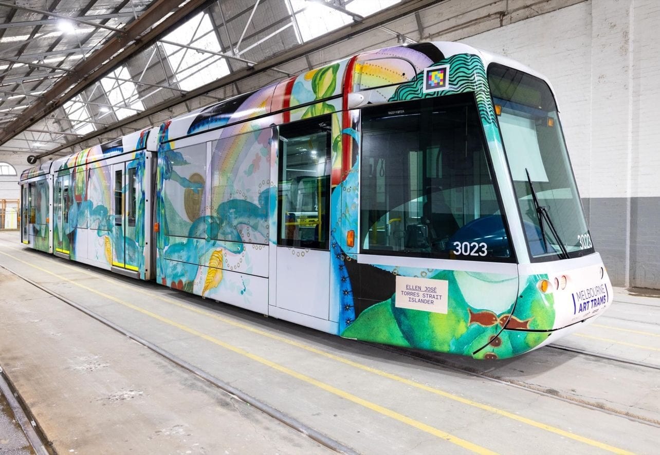 Three-quarter view of C-Class 3023 «Art Tram» inside the white depot, with yellow floor lines and maintenance tracks; the Ellen José wrap shows green human figures with botanical motifs and, above the right-hand cab, the white-framed NaviLens code next to the «MELBOURNE ART TRAMS» stamp