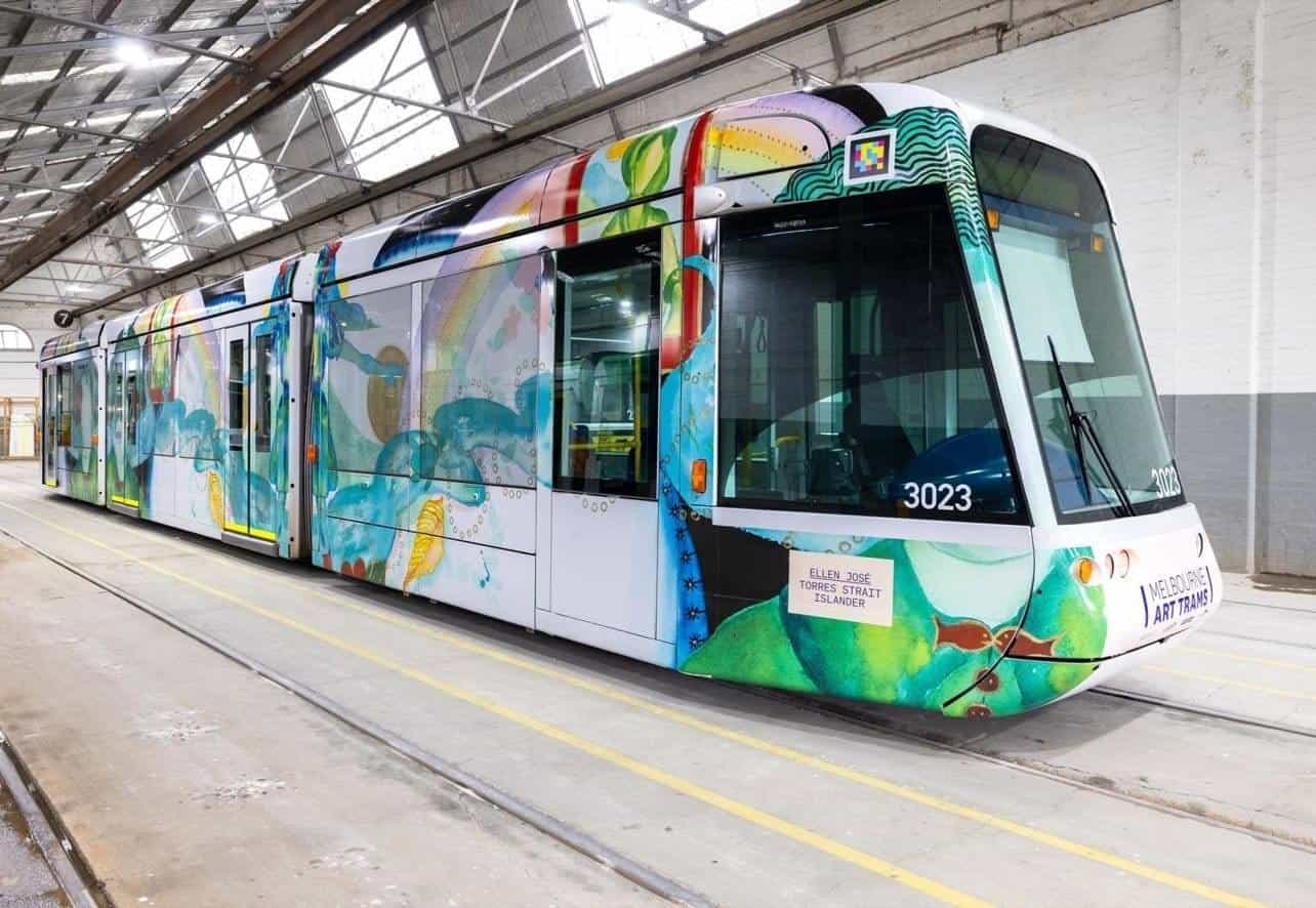 Three-quarter view of C-Class 3023 “Art Tram” inside the white depot, with yellow floor lines and maintenance tracks; the Ellen José wrap shows green human figures with botanical motifs and, above the right-hand cab, the white-framed NaviLens code next to the “MELBOURNE ART TRAMS” stamp