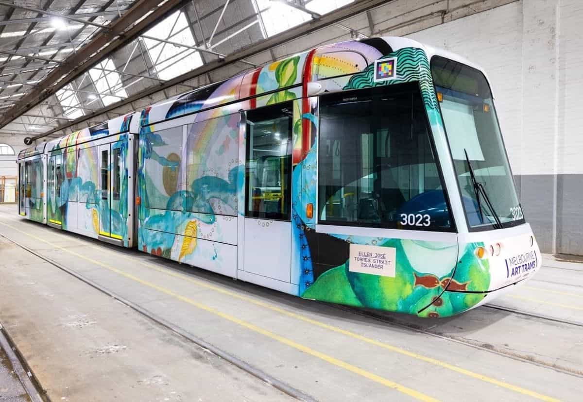 Vista en tres cuartos del C-Class 3023 «Art Tram» dentro del depósito blanco, con líneas amarillas en el suelo y vías de mantenimiento; el wrap de Ellen José muestra figuras humanas verdes con motivos botánicos y, sobre la cabina derecha, el código NaviLens enmarcado en blanco junto al sello «MELBOURNE ART TRAMS»