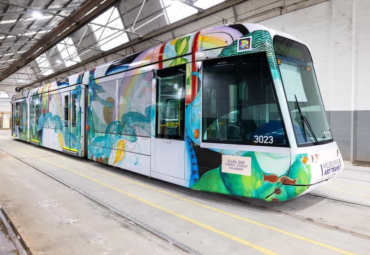 白い車庫内の C-Class 3023「Art Tram」を斜めから見た写真。床には黄色いラインと整備用線路、Ellen José のラッピングには植物モチーフを伴う緑の人物像、右側運転台の上に白枠の NaviLens コードと「MELBOURNE ART TRAMS」の刻印
