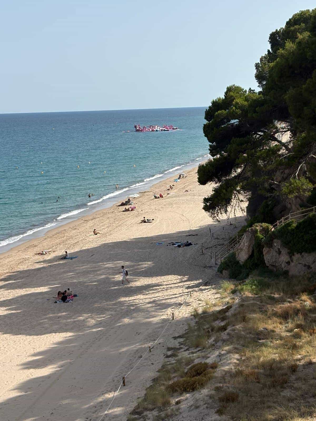 Stretch of sand along the Passeig de les Cales with an inflatable water park moored offshore and a roped path through the dunes