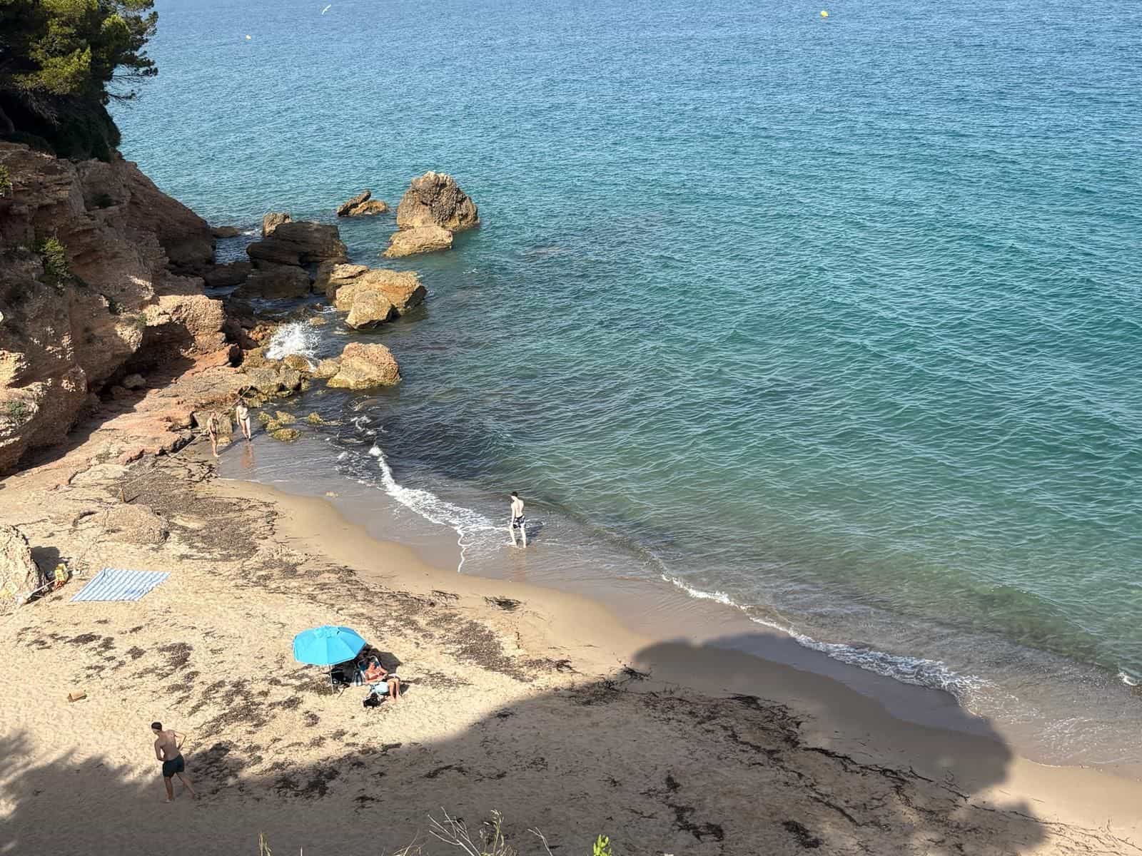 Kleine Sandbucht, umrahmt von rötlichen Felsen und türkisfarbenem Wasser, mit blauem Sonnenschirm und Badegästen am Ufer