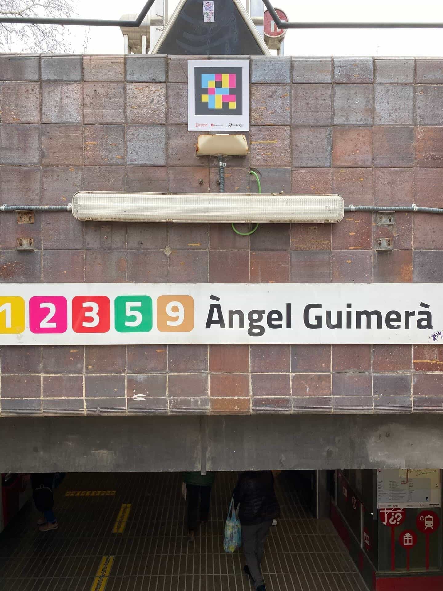 Primo piano del cartello d’accesso «1 2 3 5 9 Àngel Guimerà» con i pittogrammi di linea a colori —1 giallo, 2 magenta, 3 rosso, 5 verde, 9 ocra— e, sulla ceramica marrone, un codice NaviLens quadrato a colori con la firma di Generalitat Valenciana · Metrovalencia · NaviLens GO