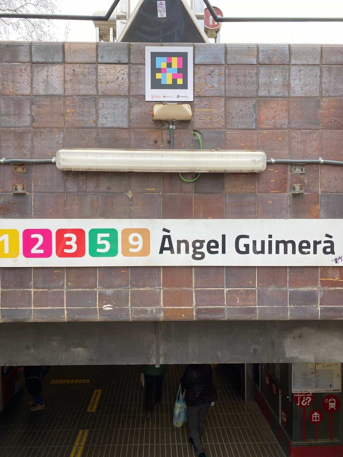 Close-up of the entrance sign '1 2 3 5 9 Àngel Guimerà' with the line pictograms in colour – 1 yellow, 2 magenta, 3 red, 5 green, 9 ochre – and, on the brown ceramic, a colourful square NaviLens code with the Generalitat Valenciana · Metrovalencia · NaviLens GO branding.
