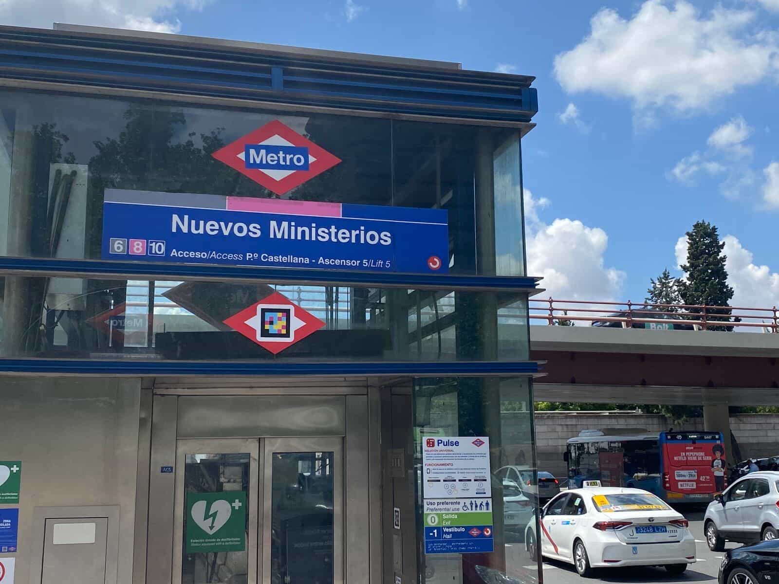 Nahaufnahme des Außenschildes von Nuevos Ministerios der Metro de Madrid: rote Metro-Raute, blaues Schild „Nuevos Ministerios“ mit den Symbolen der Linien 6, 8 und 10 und Hinweis „Zugang P.º Castellana – Aufzug 5“; neben der Raute eine identische rote Raute mit einem quadratischen, farbigen NaviLens-Code im Inneren
