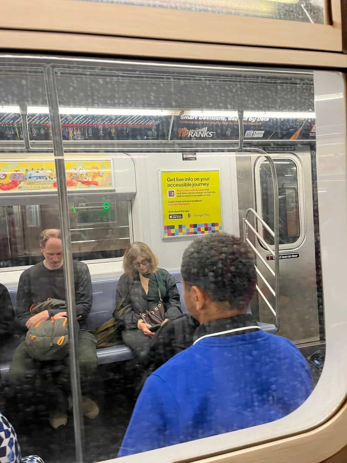 Vista desde el andén, a través de la ventanilla de un vagón R211, del mismo cartel oficial amarillo de la MTA explicando NaviLens; al fondo, viajeros sentados en el banco corrido de aluminio del coche nuevo