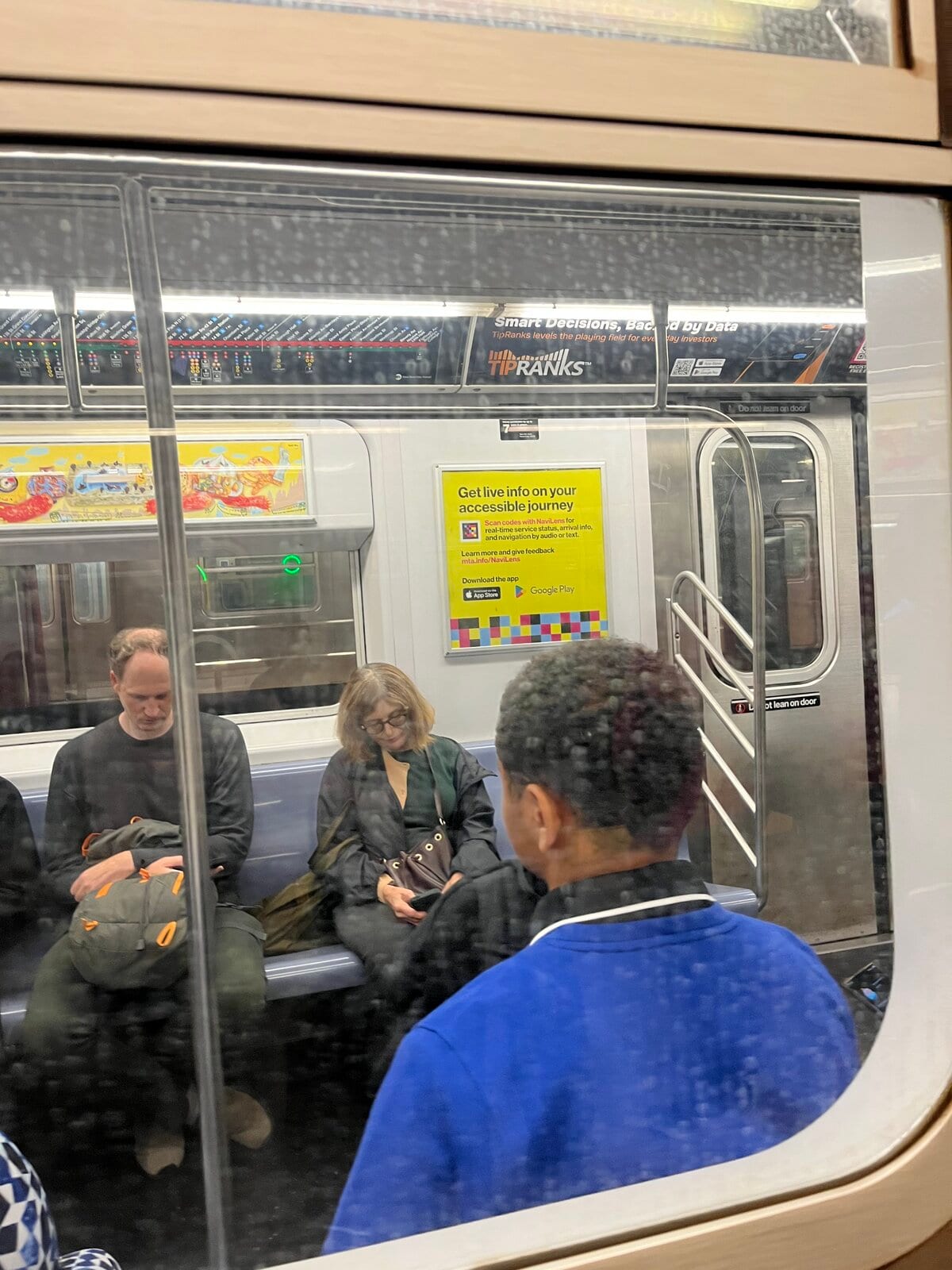 Vue depuis le quai, à travers la fenêtre d'une voiture R211, du même panneau officiel jaune de la MTA sur NaviLens ; en arrière-plan, des voyageurs assis sur le banc en aluminium de la nouvelle voiture