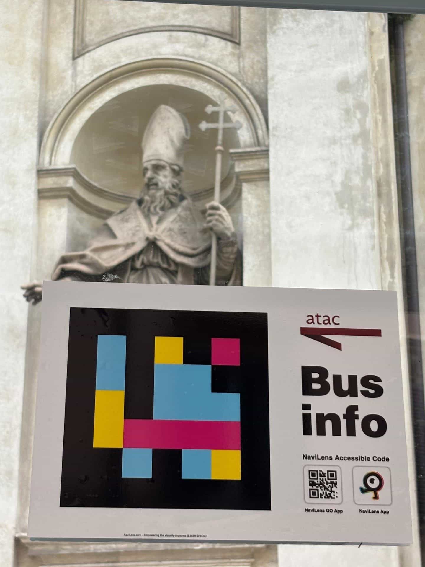 Dettaglio del cartello Bus info di ATAC con codice NaviLens accessibile e le icone di download di NaviLens GO e NaviLens App, sul vetro di una pensilina del centro di Roma