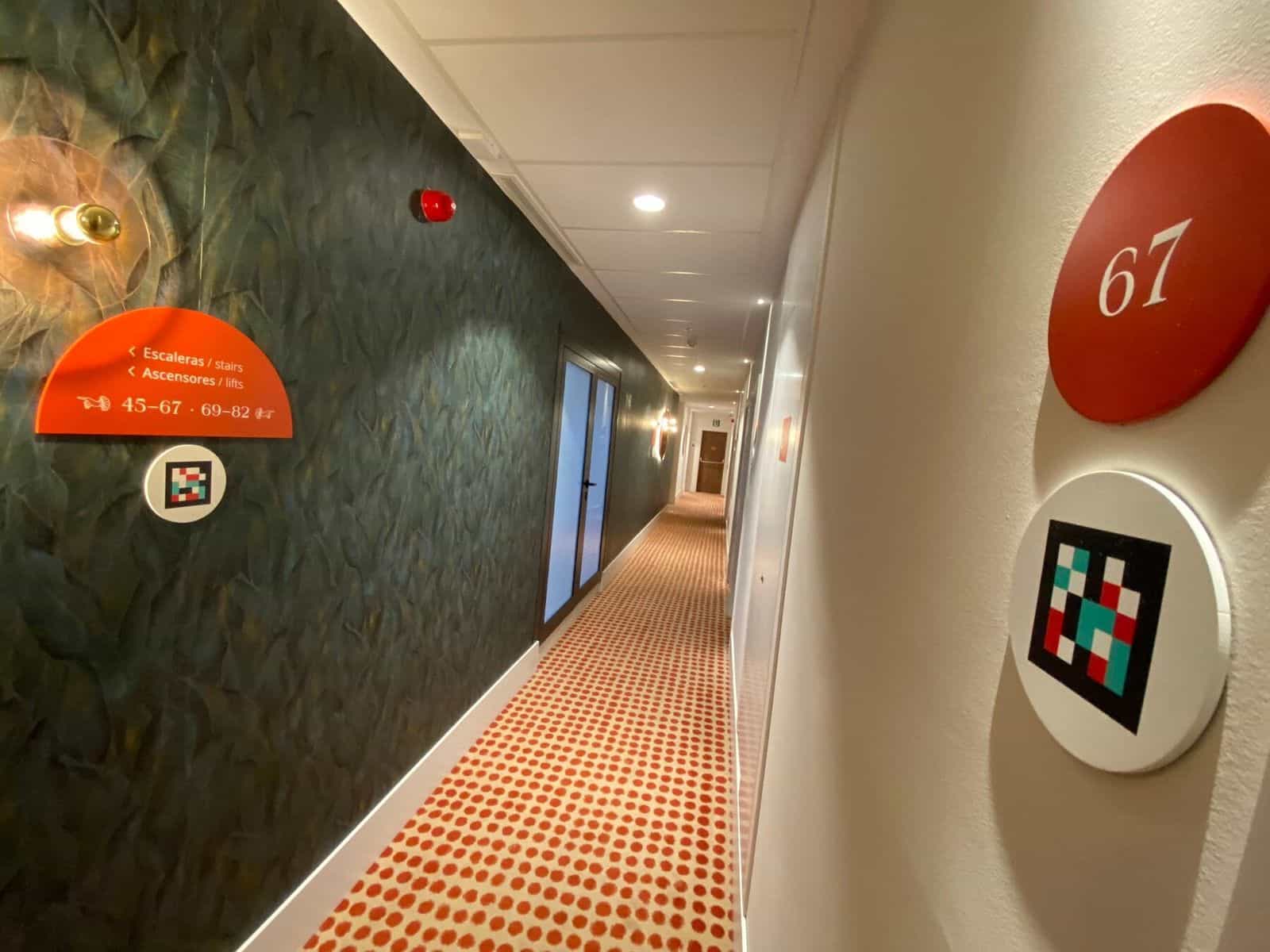 Wegweiser am ibis Styles Sevilla auf grüner Tapete: oranger Halbmond mit Linkspfeil „< Treppen · < Aufzüge · 45-67 · 69-82“ und weißer Disc mit NaviLens-Code