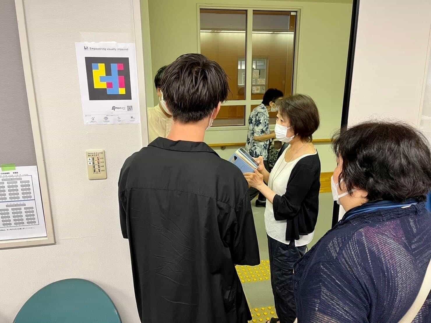 廊下の壁に貼られた、NaviLensコードとダウンロードQR付きA4「Empowering visually impaired」案内板